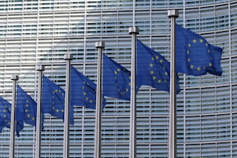 Several blue flags featuring yellow stars arranged in a circle, representing the European Union, are displayed in a row against the backdrop of a modern architectural facade with glass and metal elements.