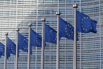 Several blue flags featuring yellow stars arranged in a circle, representing the European Union, are displayed in a row against the backdrop of a modern architectural facade with glass and metal elements.