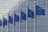 Several blue flags featuring yellow stars arranged in a circle, representing the European Union, are displayed in a row against the backdrop of a modern architectural facade with glass and metal elements.