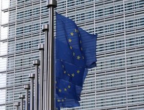 Several European Union flags with blue backgrounds and yellow stars are mounted on metal poles in front of a modern building with a grid-like facade.