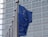 Several European Union flags with blue backgrounds and yellow stars are mounted on metal poles in front of a modern building with a grid-like facade.