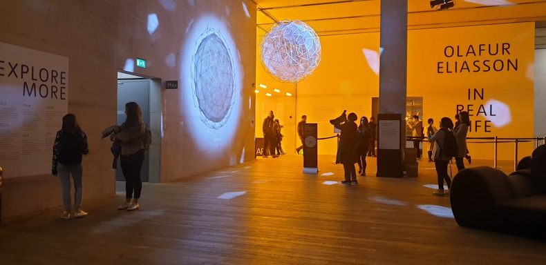 A modern exhibition hall with warm lighting, displaying artistic installations. People are observing the artwork and reading informational boards. The space has a large spherical structure hanging from the ceiling and various signs on the walls.