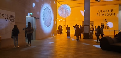 A modern exhibition hall with warm lighting, displaying artistic installations. People are observing the artwork and reading informational boards. The space has a large spherical structure hanging from the ceiling and various signs on the walls.