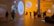 A modern exhibition hall with warm lighting, displaying artistic installations. People are observing the artwork and reading informational boards. The space has a large spherical structure hanging from the ceiling and various signs on the walls.