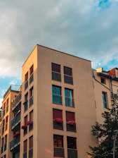 A modern apartment building with a light-colored facade features multiple balconies adorned with red flowers. The structure stands against a backdrop of a partly cloudy sky, with the golden hour sunlight casting a warm glow on its walls.