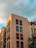 A modern apartment building with a light-colored facade features multiple balconies adorned with red flowers. The structure stands against a backdrop of a partly cloudy sky, with the golden hour sunlight casting a warm glow on its walls.