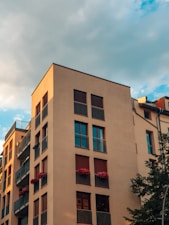 A modern apartment building with a light-colored facade features multiple balconies adorned with red flowers. The structure stands against a backdrop of a partly cloudy sky, with the golden hour sunlight casting a warm glow on its walls.