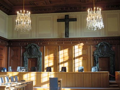An elegantly furnished courtroom is adorned with two grand chandeliers hanging from a coffered ceiling. The room's walls sport large wooden panels with decorative carvings and a prominent cross mounted above the central area. Sunlight streams through the windows, casting shadows on the polished wooden furniture and green marble features surrounding the doors at either end of the room.