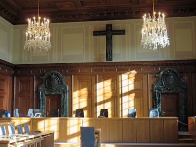 An elegantly furnished courtroom is adorned with two grand chandeliers hanging from a coffered ceiling. The room's walls sport large wooden panels with decorative carvings and a prominent cross mounted above the central area. Sunlight streams through the windows, casting shadows on the polished wooden furniture and green marble features surrounding the doors at either end of the room.