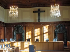 An elegantly furnished courtroom is adorned with two grand chandeliers hanging from a coffered ceiling. The room's walls sport large wooden panels with decorative carvings and a prominent cross mounted above the central area. Sunlight streams through the windows, casting shadows on the polished wooden furniture and green marble features surrounding the doors at either end of the room.