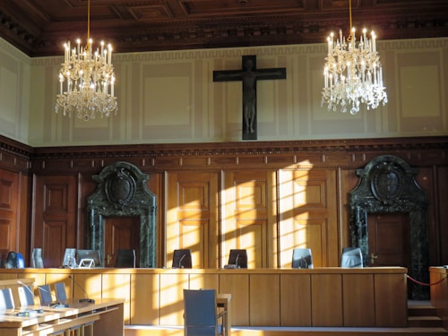 An elegantly furnished courtroom is adorned with two grand chandeliers hanging from a coffered ceiling. The room's walls sport large wooden panels with decorative carvings and a prominent cross mounted above the central area. Sunlight streams through the windows, casting shadows on the polished wooden furniture and green marble features surrounding the doors at either end of the room.