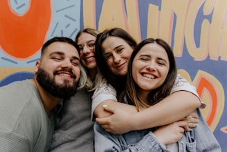 A group of four people smiling and embracing each other in front of a colorful mural with bold, abstract shapes and letters. The individuals appear joyful and close, with two of them wearing glasses. The background is vibrant with shades of orange, blue, and yellow.