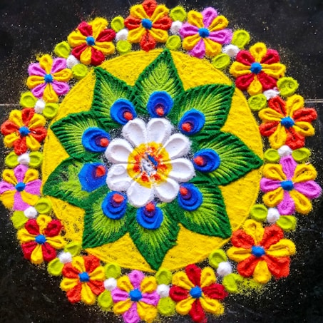 A vibrant and intricate rangoli pattern featuring a central flower surrounded by layered designs. The artwork includes a white flower with blue peacock-shaped designs and green leaves on a bright yellow background. Surrounding the central pattern is a border of colorful flowers made of petals in red, yellow, pink, and purple, interspersed with small green circles.