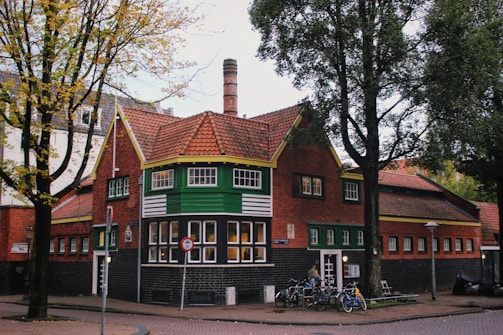 A quaint brick building with a red-tiled roof and green accents is surrounded by trees with autumn leaves. Bicycles are parked near the entrance, and a tall streetlamp stands nearby. The architecture features a mix of traditional and rustic styles, with large windows and wooden details.