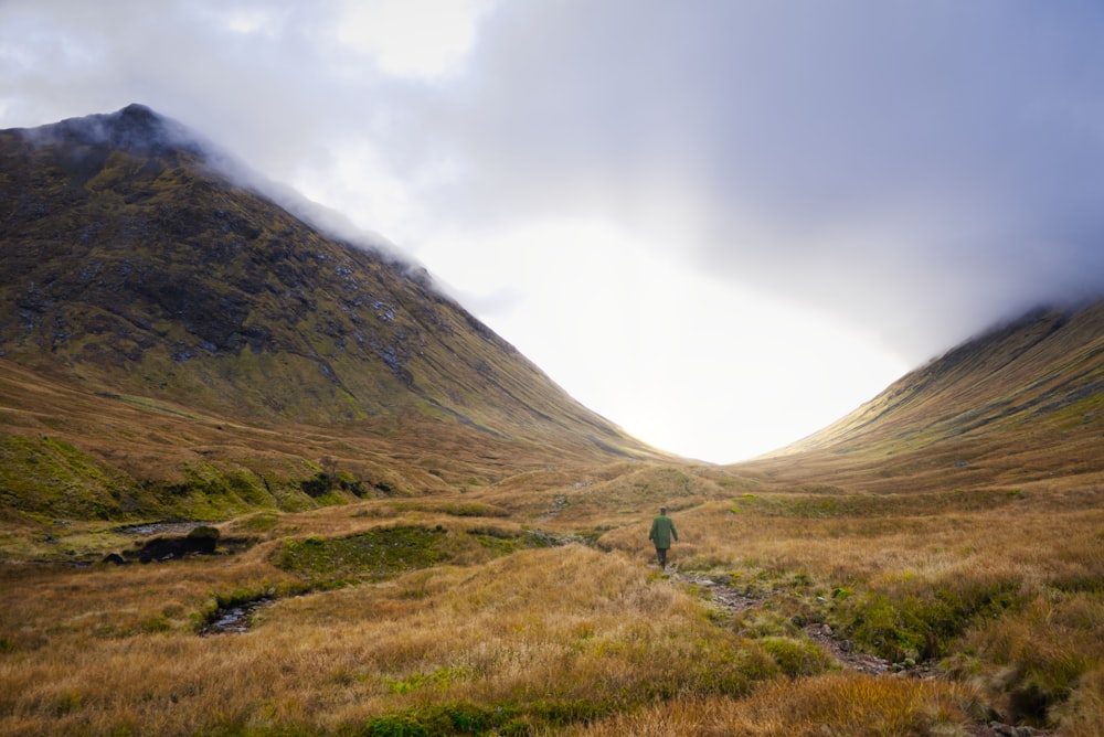 person in green jacket walking on green grass field near mountain during daytime