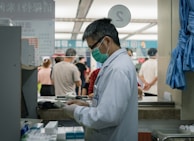 A healthcare professional wearing a white coat and a green face mask is standing at a counter or a pharmacy. They are focused on a piece of paper or prescription pad. Behind them, several people are waiting in line, suggesting a public or clinical setting. Various medical supplies and garments are visible around the area.