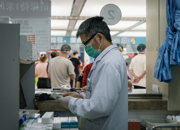 A healthcare professional wearing a white coat and a green face mask is standing at a counter or a pharmacy. They are focused on a piece of paper or prescription pad. Behind them, several people are waiting in line, suggesting a public or clinical setting. Various medical supplies and garments are visible around the area.