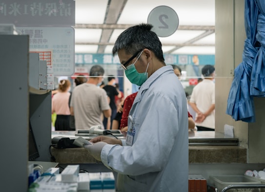 A healthcare professional wearing a white coat and a green face mask is standing at a counter or a pharmacy. They are focused on a piece of paper or prescription pad. Behind them, several people are waiting in line, suggesting a public or clinical setting. Various medical supplies and garments are visible around the area.