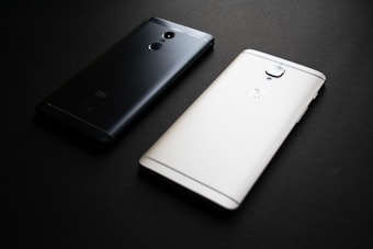 Two smartphones placed on a dark surface, one with a black back and the other with a white back. The black phone has a brand logo and camera feature visible, while the white phone also displays a similar logo and camera arrangement.