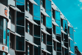 An urban building with mirrored windows reflecting the sky and surrounding structures. The detailed architecture includes angular window shades protruding from the facade. There is a sign with the word 'Lira' and a stylized logo on the left side.