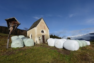 A small, rustic chapel with a steep roof stands in a grassy field. Wrapped hay bales are positioned around the chapel. To the left, a wooden crucifix with a figure of Jesus is visible. The background includes sparse forestation and a distant mountain range partially obscured by clouds.