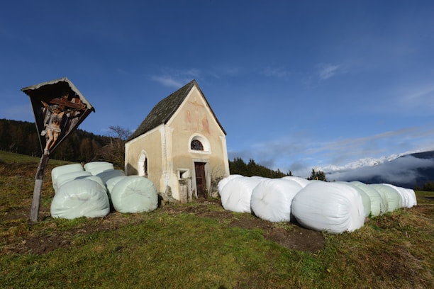 A small, rustic chapel with a steep roof stands in a grassy field. Wrapped hay bales are positioned around the chapel. To the left, a wooden crucifix with a figure of Jesus is visible. The background includes sparse forestation and a distant mountain range partially obscured by clouds.