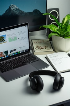 A neatly organized desk features a laptop displaying a webpage, a potted plant, a pair of black headphones, a notebook with notes and sketches, and a book titled '100 Startup'. In the background, a larger monitor shows a serene mountain landscape with a starry sky. The desk has a minimalist and modern aesthetic.