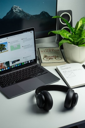 A neatly organized desk features a laptop displaying a webpage, a potted plant, a pair of black headphones, a notebook with notes and sketches, and a book titled '100 Startup'. In the background, a larger monitor shows a serene mountain landscape with a starry sky. The desk has a minimalist and modern aesthetic.