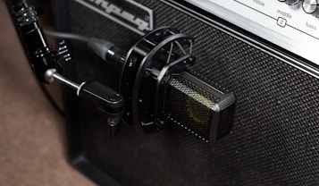 A close-up view of a professional microphone positioned in front of an amplifier. The microphone is securely attached to a stand, pointing towards the speaker area of the amplifier. The amplifier features a textured surface and control panel with labels for treble, middle, and bass. A cable connects the microphone, indicating a setup for capturing sound.
