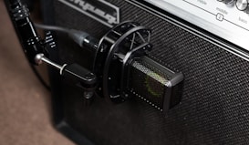 A close-up view of a professional microphone positioned in front of an amplifier. The microphone is securely attached to a stand, pointing towards the speaker area of the amplifier. The amplifier features a textured surface and control panel with labels for treble, middle, and bass. A cable connects the microphone, indicating a setup for capturing sound.