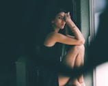 A person sits by a window, resting their head on their knees with an introspective expression. The lighting creates soft shadows on their face and highlights the textures of their dark clothing and jewelry.