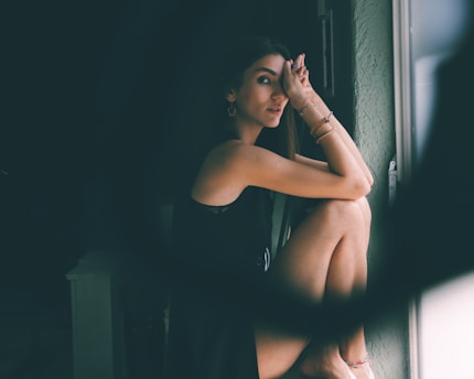 A person sits by a window, resting their head on their knees with an introspective expression. The lighting creates soft shadows on their face and highlights the textures of their dark clothing and jewelry.