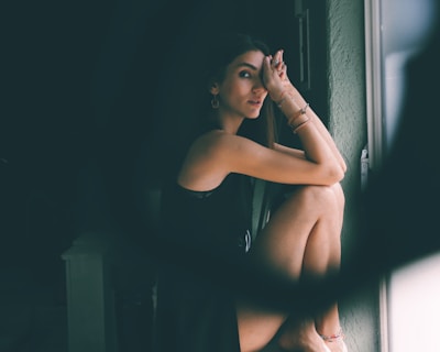 A person sits by a window, resting their head on their knees with an introspective expression. The lighting creates soft shadows on their face and highlights the textures of their dark clothing and jewelry.