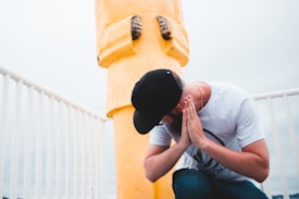 A person wearing a black cap and glasses is kneeling and holding their hands together in a praying gesture. They are positioned next to a large, yellow statue that appears to be a figure from the chest down. The setting includes a white fence and an overcast sky.