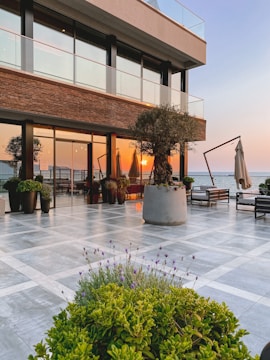 A luxurious outdoor patio area by the sea with modern furniture and large potted plants. The sun is setting, casting a warm glow over the scene. The building has a contemporary design with glass railings and brick accents.