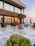 A luxurious outdoor patio area by the sea with modern furniture and large potted plants. The sun is setting, casting a warm glow over the scene. The building has a contemporary design with glass railings and brick accents.