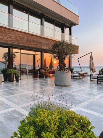 A luxurious outdoor patio area by the sea with modern furniture and large potted plants. The sun is setting, casting a warm glow over the scene. The building has a contemporary design with glass railings and brick accents.