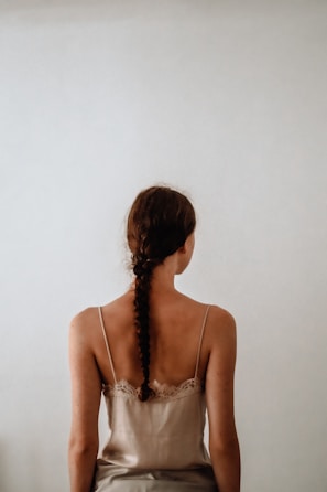 A woman with a braided hairstyle sits with her back to the viewer, wearing a spaghetti strap top. The setting is minimalistic with a plain, light-colored background.