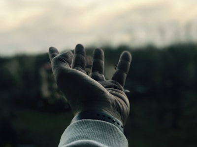 A close-up of an open hand reaching out against a blurred background of moody, overcast skies and faint outlines of trees. The hand is slightly tilted with fingers softly curled, wearing a light-colored sleeve and a wristband.