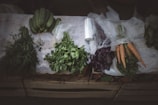 A variety of fresh vegetables are neatly arranged on a table, including cucumbers, carrots, and different herbs. The table is covered with a lace cloth, and some of the vegetables are wrapped in plastic. The setting has a rustic feel with a subdued lighting enhancing the freshness and colors of the produce.