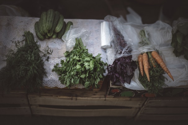 A variety of fresh vegetables are neatly arranged on a table, including cucumbers, carrots, and different herbs. The table is covered with a lace cloth, and some of the vegetables are wrapped in plastic. The setting has a rustic feel with a subdued lighting enhancing the freshness and colors of the produce.