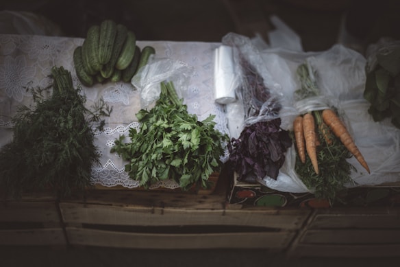 A variety of fresh vegetables are neatly arranged on a table, including cucumbers, carrots, and different herbs. The table is covered with a lace cloth, and some of the vegetables are wrapped in plastic. The setting has a rustic feel with a subdued lighting enhancing the freshness and colors of the produce.