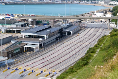 A large transportation terminal with multiple lanes for vehicles arranged in parallel stretches across the scene. Nearby, several buildings with solar panels on their roofs are visible. In the background, a body of water and parts of a coastal town are seen. The area appears organized and structured, with clear lanes and signage.