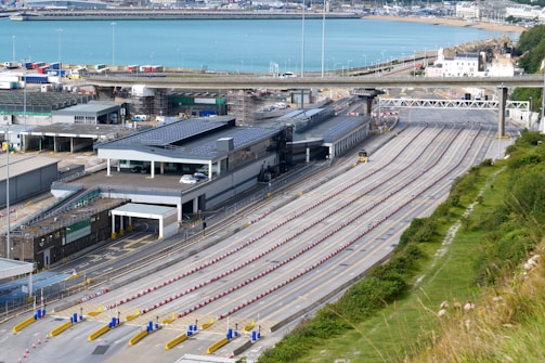 A large transportation terminal with multiple lanes for vehicles arranged in parallel stretches across the scene. Nearby, several buildings with solar panels on their roofs are visible. In the background, a body of water and parts of a coastal town are seen. The area appears organized and structured, with clear lanes and signage.