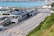 A large transportation terminal with multiple lanes for vehicles arranged in parallel stretches across the scene. Nearby, several buildings with solar panels on their roofs are visible. In the background, a body of water and parts of a coastal town are seen. The area appears organized and structured, with clear lanes and signage.