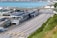 A large transportation terminal with multiple lanes for vehicles arranged in parallel stretches across the scene. Nearby, several buildings with solar panels on their roofs are visible. In the background, a body of water and parts of a coastal town are seen. The area appears organized and structured, with clear lanes and signage.