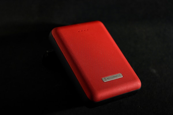 A red, textured rectangular electronic device with a labeled metallic plate on one side is positioned against a dark background. The upper side of the device has four small LED lights.