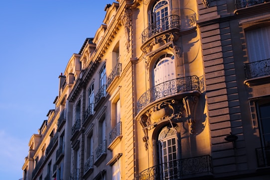 The photograph captures the facade of a European-style building bathed in warm sunlight, showing intricate iron balconies and detailed stonework with tall, narrow windows that highlight the architectural elegance.