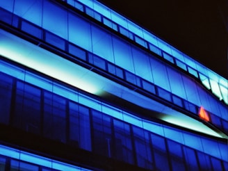 A modern architectural structure with large glass panels illuminated by vibrant blue lights. The building features sleek lines and dark shadows, creating a futuristic and dynamic aesthetic.