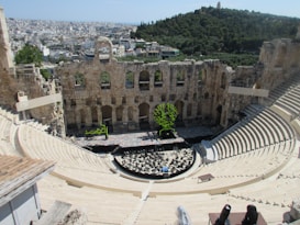 An ancient amphitheater with stone seating arranged in a semi-circular fashion. The stage area features modern installations, including abstract art pieces. A cityscape with densely packed buildings extends into the background, while a green hill provides contrast.